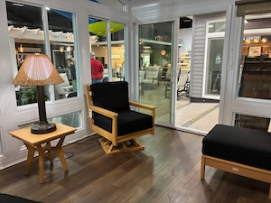 An indoor showroom display featuring a cozy sunroom or patio enclosure setup. A wooden armchair with thick black cushions sits on dark wood-plank flooring next to a matching end table with a traditional lamp. To the right, a matching black cushioned ottoman is visible. Large windows and a sliding glass door reveal more furniture displays in the background, creating a bright and inviting atmosphere.