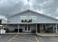 An exterior shot of Raber Patio Enclosures & Furniture, LLC, a light blue, gabled storefront under a cloudy sky. A large sign with green and black text is mounted on the front, featuring silhouettes of Adirondack chairs. The porch is filled with outdoor furniture displays, including cushioned sofas, wooden chairs, and tables. To the left, the words "RELAX," "RENEW," and "REFRESH" are painted on the wall above a display area, and a blue ADA-compliant parking sign is visible in the foreground.