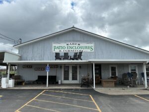 An exterior shot of Raber Patio Enclosures & Furniture, LLC, a light blue, gabled storefront under a cloudy sky. A large sign with green and black text is mounted on the front, featuring silhouettes of Adirondack chairs. The porch is filled with outdoor furniture displays, including cushioned sofas, wooden chairs, and tables. To the left, the words "RELAX," "RENEW," and "REFRESH" are painted on the wall above a display area, and a blue ADA-compliant parking sign is visible in the foreground.
