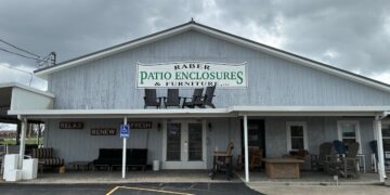 An exterior shot of Raber Patio Enclosures & Furniture, LLC, a light blue, gabled storefront under a cloudy sky. A large sign with green and black text is mounted on the front, featuring silhouettes of Adirondack chairs. The porch is filled with outdoor furniture displays, including cushioned sofas, wooden chairs, and tables. To the left, the words "RELAX," "RENEW," and "REFRESH" are painted on the wall above a display area, and a blue ADA-compliant parking sign is visible in the foreground.