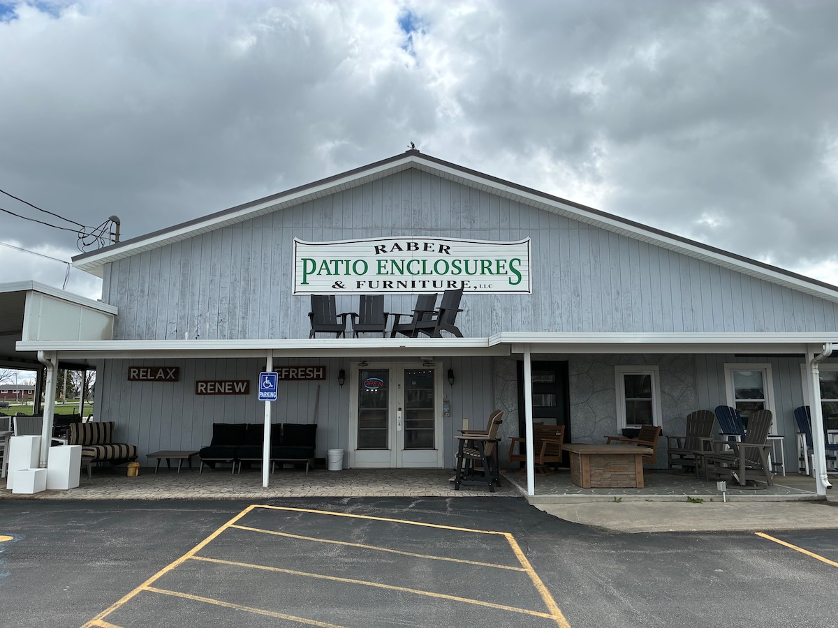 An exterior shot of Raber Patio Enclosures & Furniture, LLC, a light blue, gabled storefront under a cloudy sky. A large sign with green and black text is mounted on the front, featuring silhouettes of Adirondack chairs. The porch is filled with outdoor furniture displays, including cushioned sofas, wooden chairs, and tables. To the left, the words "RELAX," "RENEW," and "REFRESH" are painted on the wall above a display area, and a blue ADA-compliant parking sign is visible in the foreground.
