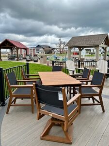 An outdoor display area at an equipment and furniture dealership features a rectangular wooden dining table surrounded by six matching armchairs with black mesh seats and backs. The set is arranged on a light grey deck. In the background, various small wooden structures, including a red-roofed pavilion and a tan shingled gazebo, are scattered across a grassy lot under a dramatic, cloudy sky.