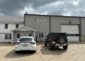 The exterior of D.A. Hochstetler & Sons, a large metal industrial building with a tan corrugated facade and a white gabled entrance. A white car and a dark red Jeep Wrangler are parked on the concrete lot in front of the building. The sky above is overcast with heavy grey clouds.