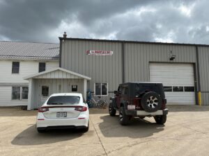 The exterior of D.A. Hochstetler & Sons, a large metal industrial building with a tan corrugated facade and a white gabled entrance. A white car and a dark red Jeep Wrangler are parked on the concrete lot in front of the building. The sky above is overcast with heavy grey clouds.