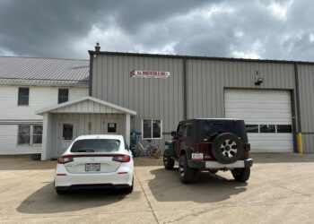 The exterior of D.A. Hochstetler & Sons, a large metal industrial building with a tan corrugated facade and a white gabled entrance. A white car and a dark red Jeep Wrangler are parked on the concrete lot in front of the building. The sky above is overcast with heavy grey clouds.