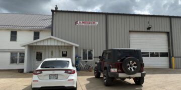 The exterior of D.A. Hochstetler & Sons, a large metal industrial building with a tan corrugated facade and a white gabled entrance. A white car and a dark red Jeep Wrangler are parked on the concrete lot in front of the building. The sky above is overcast with heavy grey clouds.