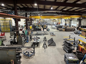 The wide-angle interior view of D.A. Hochstetler & Sons' manufacturing facility in Topeka, Indiana. The large shop floor features various pieces of heavy-duty machinery, industrial workbenches, and steel components for products like StackPro racking and Reliable attachments. Several yellow overhead crane systems span the ceiling above the workstations.