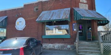 An exterior shot of The Rustic Spoon restaurant, a red brick building with a stone foundation and vintage charm. The facade features corrugated metal awnings over the windows and a green-and-white striped awning over the entrance. A circular white logo for the restaurant is mounted on the brick wall to the left of the main window, which has an "OPEN" neon sign. A plaque on the corner of the building reads "EMMA 1930." A black Cadillac is parked in the foreground.