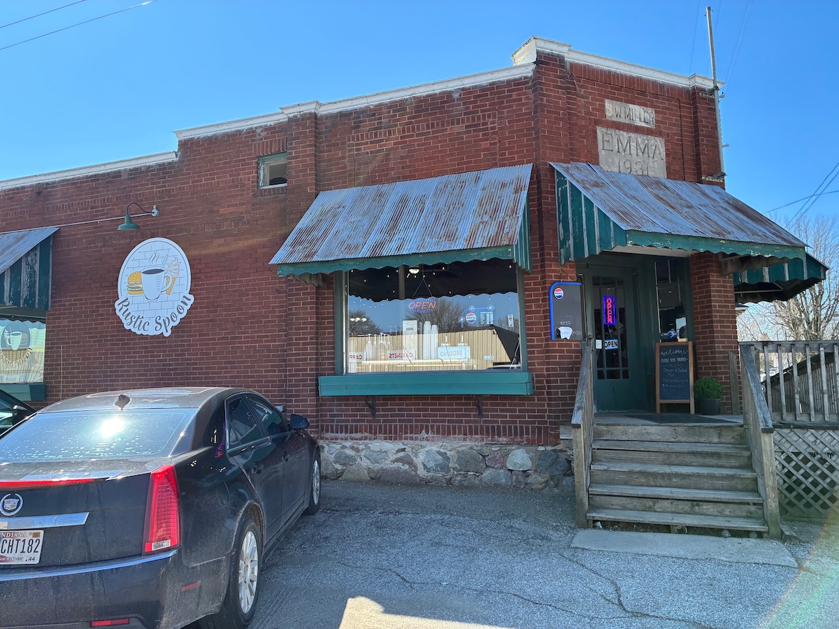 An exterior shot of The Rustic Spoon restaurant, a red brick building with a stone foundation and vintage charm. The facade features corrugated metal awnings over the windows and a green-and-white striped awning over the entrance. A circular white logo for the restaurant is mounted on the brick wall to the left of the main window, which has an "OPEN" neon sign. A plaque on the corner of the building reads "EMMA 1930." A black Cadillac is parked in the foreground.