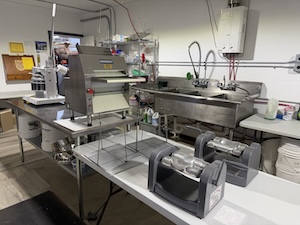 An indoor commercial kitchen or production space featuring stainless steel equipment. On a metal prep table, there is a specialized dough sheeter or roller machine. To the right is a large three-compartment industrial sink with a high-neck faucet. In the foreground, two grey mechanical tape dispensers sit on a white folding table, suggesting a space used for both food preparation and packaging.