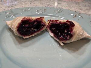 A flaky, golden-brown hand pie is broken in half on a light blue decorative plate, revealing a thick, dark purple blueberry filling. The glossy fruit filling oozes slightly from the buttery, crimped crust. The plate has embossed nautical designs, and the background shows a neutral-toned speckled countertop.