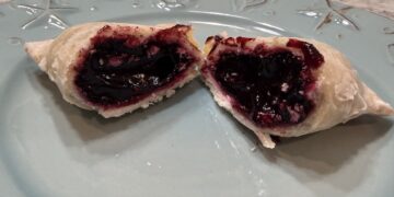 A flaky, golden-brown hand pie is broken in half on a light blue decorative plate, revealing a thick, dark purple blueberry filling. The glossy fruit filling oozes slightly from the buttery, crimped crust. The plate has embossed nautical designs, and the background shows a neutral-toned speckled countertop.