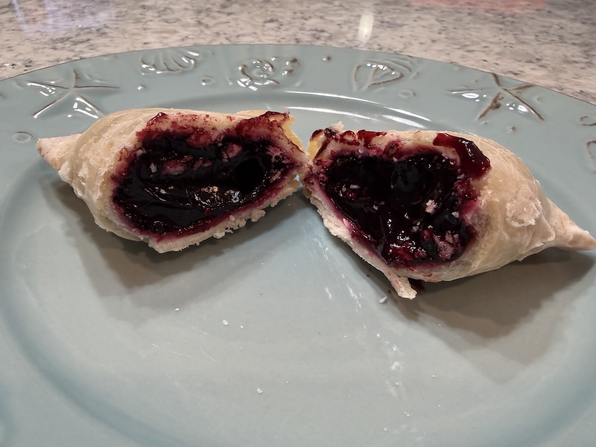 A flaky, golden-brown hand pie is broken in half on a light blue decorative plate, revealing a thick, dark purple blueberry filling. The glossy fruit filling oozes slightly from the buttery, crimped crust. The plate has embossed nautical designs, and the background shows a neutral-toned speckled countertop.