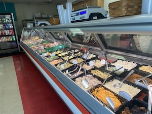 A long, glass-fronted deli display case inside Kingsley Meats & Catering. The case is filled with over a dozen black rectangular trays containing various prepared salads and side dishes, including potato salad, pasta salads, and pimento cheese. Each tray has a serving spoon or tongs. In the background, a digital scale sits on the counter, and a refrigerated beverage case is visible against the wall. The floor has a distinctive red stripe running parallel to the counter.