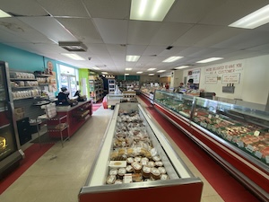 An expansive view of the retail floor at Kingsley Meats & Catering in Louisville, KY. The shop features long, brightly lit refrigerated display cases running parallel through the center and right side of the room, filled with fresh cuts of meat and gourmet deli sides. To the left, a checkout counter sits against a teal-colored wall lined with shelves of specialty seasonings and sauces. The white walls are adorned with customer service signs, and the space has a clean, organized, old-fashioned butcher shop feel.