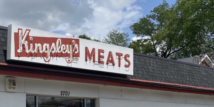 The storefront of Kingsley Meats at 2701 Taylorsville Road. A large white sign with red lettering reads "Kingsley's MEATS," with the first word in a stylized script. Below it, the glass entrance doors are marked with the address "2701." To the right, on the white building wall, another sign reads "Kingsley's AGED PRIME BEEF" in red. The sky above is bright blue with scattered white clouds, and green treetops are visible in the background.