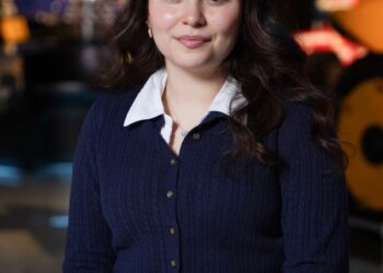 A medium-shot, eye-level portrait of a smiling young woman with long, dark, wavy hair, identified as Melanie Guterrez, a 2026 500 Festival Princess. She is wearing a navy blue cable-knit cardigan over a white collared shirt and black pants. She is seated with her hands clasped in her lap, wearing a silver charm bracelet. The background is a modern museum setting, likely the Indianapolis Motor Speedway Museum, with blurred exhibits including a large yellow racing machine on the right and a silver trophy-like structure on the left.