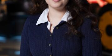 A medium-shot, eye-level portrait of a smiling young woman with long, dark, wavy hair, identified as Melanie Guterrez, a 2026 500 Festival Princess. She is wearing a navy blue cable-knit cardigan over a white collared shirt and black pants. She is seated with her hands clasped in her lap, wearing a silver charm bracelet. The background is a modern museum setting, likely the Indianapolis Motor Speedway Museum, with blurred exhibits including a large yellow racing machine on the right and a silver trophy-like structure on the left.