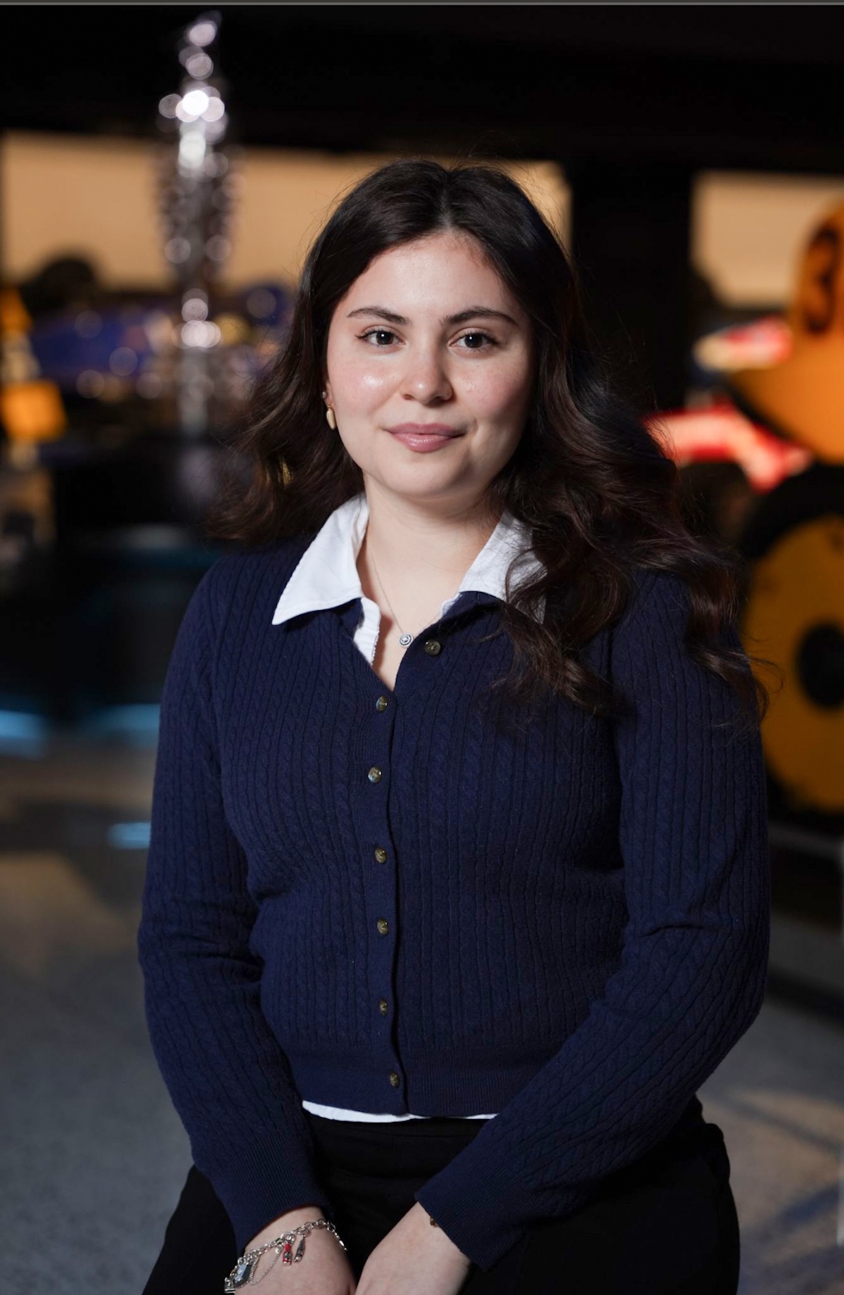 A medium-shot, eye-level portrait of a smiling young woman with long, dark, wavy hair, identified as Melanie Guterrez, a 2026 500 Festival Princess. She is wearing a navy blue cable-knit cardigan over a white collared shirt and black pants. She is seated with her hands clasped in her lap, wearing a silver charm bracelet. The background is a modern museum setting, likely the Indianapolis Motor Speedway Museum, with blurred exhibits including a large yellow racing machine on the right and a silver trophy-like structure on the left.