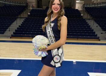 A smiling young woman with long wavy light brown hair stands on a basketball court, wearing a navy blue dress and a sparkling silver tiara. She wears a white sash with "500 FESTIVAL PRINCESS" printed in black letters and holds silver and iridescent pom-poms in her right hand. Behind her, the wall is lined with championship banners, including a prominent one that reads "TRINE MEN'S BASKETBALL NCAA NATIONAL CHAMPIONS 2024."