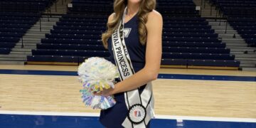 A smiling young woman with long wavy light brown hair stands on a basketball court, wearing a navy blue dress and a sparkling silver tiara. She wears a white sash with "500 FESTIVAL PRINCESS" printed in black letters and holds silver and iridescent pom-poms in her right hand. Behind her, the wall is lined with championship banners, including a prominent one that reads "TRINE MEN'S BASKETBALL NCAA NATIONAL CHAMPIONS 2024."