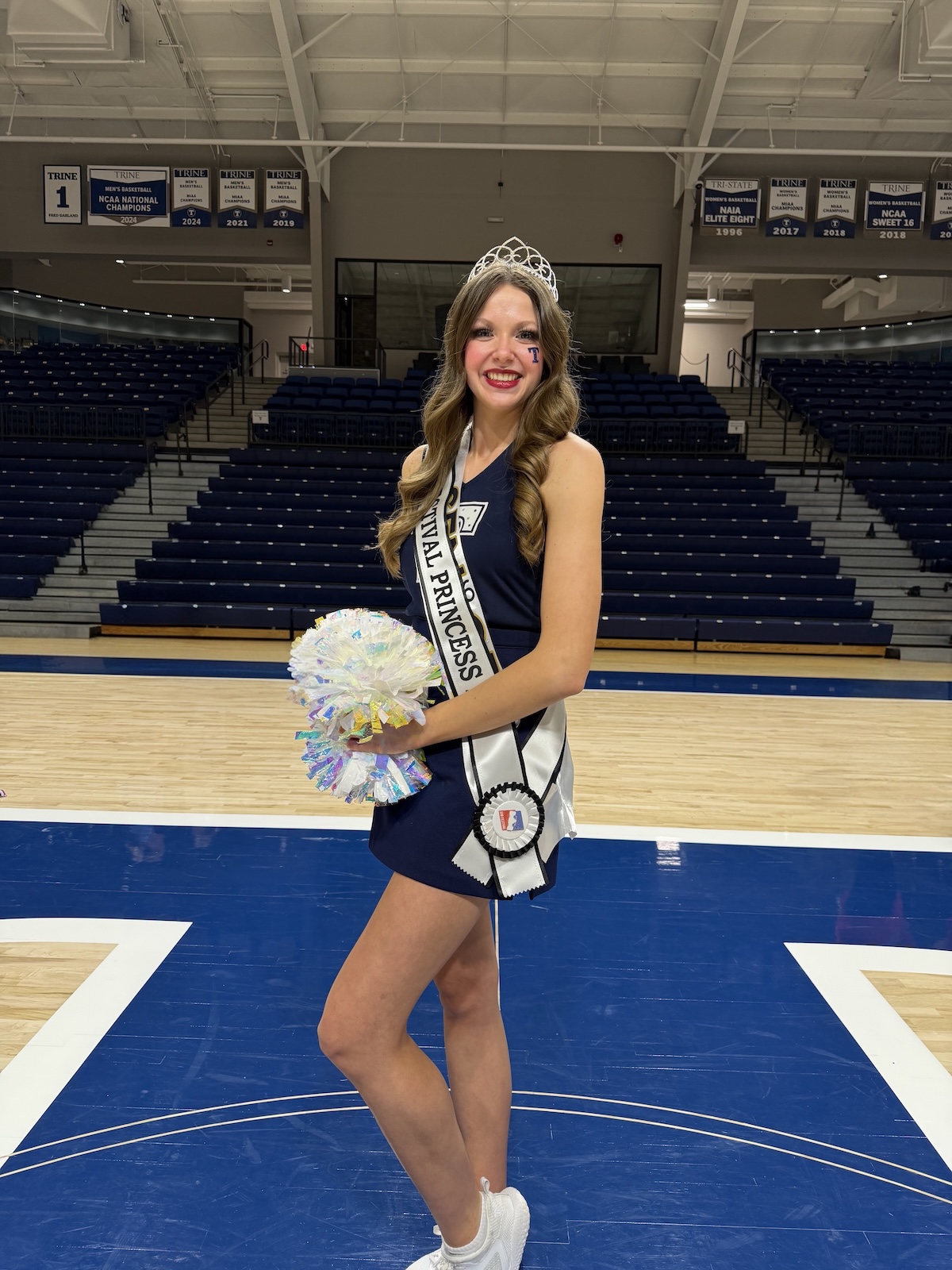 A smiling young woman with long wavy light brown hair stands on a basketball court, wearing a navy blue dress and a sparkling silver tiara. She wears a white sash with "500 FESTIVAL PRINCESS" printed in black letters and holds silver and iridescent pom-poms in her right hand. Behind her, the wall is lined with championship banners, including a prominent one that reads "TRINE MEN'S BASKETBALL NCAA NATIONAL CHAMPIONS 2024."