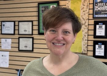 A woman with short, styled brown hair and a warm smile stands in the foreground, wearing a sage green V-neck sweater and gold hoop earrings. She is positioned in front of a wooden slat wall adorned with numerous framed awards and certificates. Notable awards include a "2021 Best of the Best" winner plaque from the Courier Journal and several "LEO Reader's Choice" certificates, suggesting a professional setting like a wellness center or office.