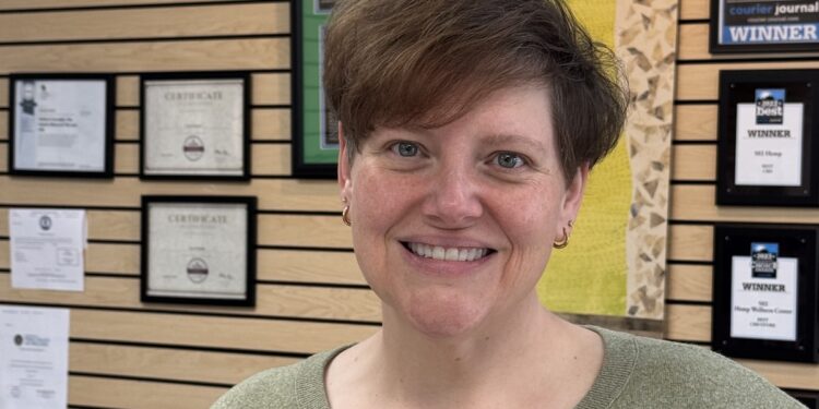 A woman with short, styled brown hair and a warm smile stands in the foreground, wearing a sage green V-neck sweater and gold hoop earrings. She is positioned in front of a wooden slat wall adorned with numerous framed awards and certificates. Notable awards include a "2021 Best of the Best" winner plaque from the Courier Journal and several "LEO Reader's Choice" certificates, suggesting a professional setting like a wellness center or office.
