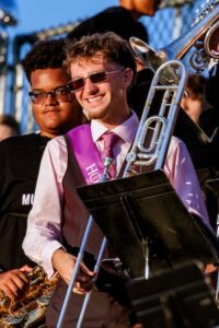 A young man with light brown hair and sunglasses is smiling broadly while playing a trombone in a marching band. He is dressed in a light pink button-down shirt, a matching tie, and a purple sash that partially reads "HOPE." He is holding his trombone in front of a music stand. To his left, another student in a black t-shirt and glasses is holding a saxophone, and a chain-link fence is visible in the background under a clear, late-afternoon sky.