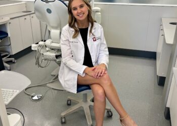 A full-shot of a smiling young woman, identified as Hannah Pensyl, a student in the Indiana University School of Dentistry Class of 2028. She is sitting in a blue and white dental chair inside a bright, modern clinic, likely the Fritts Clinical Care Center. She is wearing a black dress and a waist-length white student clinic coat featuring the IU School of Dentistry logo and her name embroidered on the front. The background shows a state-of-the-art dental facility with multiple treatment stations, medical monitors, and overhead exam lights.