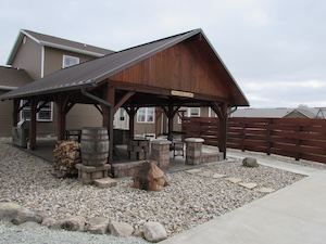 A large wooden timber-frame pavilion with a dark metal gabled roof is situated in a backyard. The pavilion features a dark-stained wood structure and sits on a stone base. Decorative elements include a large wooden barrel and a small pile of firewood. The surrounding ground is covered in light grey gravel with a concrete walkway leading to the area. A horizontal wooden privacy fence runs behind the structure, and a two-story tan house is visible in the background under an overcast sky.