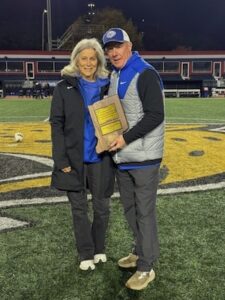 A man and a woman stand together on a football field with a large yellow and blue lion logo; the man, wearing a grey vest and a blue HSE baseball cap, holds a wooden plaque while the woman stands beside him in a black jacket.