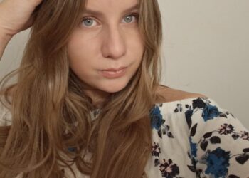 A close-up selfie of a young woman with long, light brown hair. She has blue eyes and is wearing a white off-the-shoulder top with a blue and pink floral pattern. She is looking directly at the camera with her right hand resting near the top of her head.