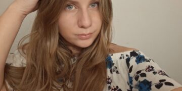 A close-up selfie of a young woman with long, light brown hair. She has blue eyes and is wearing a white off-the-shoulder top with a blue and pink floral pattern. She is looking directly at the camera with her right hand resting near the top of her head.