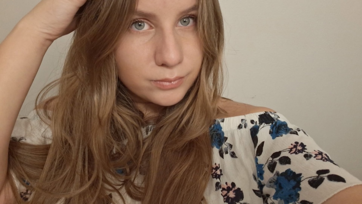 A close-up selfie of a young woman with long, light brown hair. She has blue eyes and is wearing a white off-the-shoulder top with a blue and pink floral pattern. She is looking directly at the camera with her right hand resting near the top of her head.