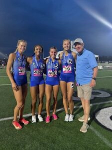 Four female athletes from the Hamilton Southeastern track team, wearing blue uniforms and medals, pose on a football field at night with Athletic Director Jim Self, who is wearing a grey vest and a blue HSE hat.