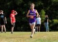 A young male cross-country runner with curly hair is captured mid-stride during a race. He is wearing a purple singlet with "MUNCIE" in white letters across the chest and a red race bib numbered "726." He is running on a grassy course toward the camera, with a focused expression. In the blurred background, several spectators stand near a line of dense green trees, including one man in a bright red shirt and hat shielding his eyes from the sun.