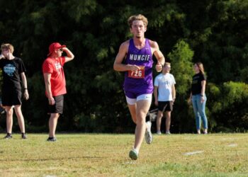 A young male cross-country runner with curly hair is captured mid-stride during a race. He is wearing a purple singlet with "MUNCIE" in white letters across the chest and a red race bib numbered "726." He is running on a grassy course toward the camera, with a focused expression. In the blurred background, several spectators stand near a line of dense green trees, including one man in a bright red shirt and hat shielding his eyes from the sun.