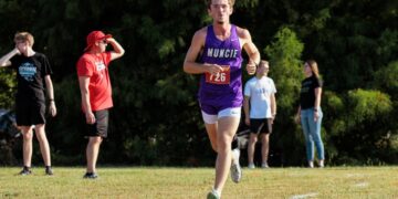 A young male cross-country runner with curly hair is captured mid-stride during a race. He is wearing a purple singlet with "MUNCIE" in white letters across the chest and a red race bib numbered "726." He is running on a grassy course toward the camera, with a focused expression. In the blurred background, several spectators stand near a line of dense green trees, including one man in a bright red shirt and hat shielding his eyes from the sun.