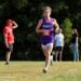A young male cross-country runner with curly hair is captured mid-stride during a race. He is wearing a purple singlet with "MUNCIE" in white letters across the chest and a red race bib numbered "726." He is running on a grassy course toward the camera, with a focused expression. In the blurred background, several spectators stand near a line of dense green trees, including one man in a bright red shirt and hat shielding his eyes from the sun.