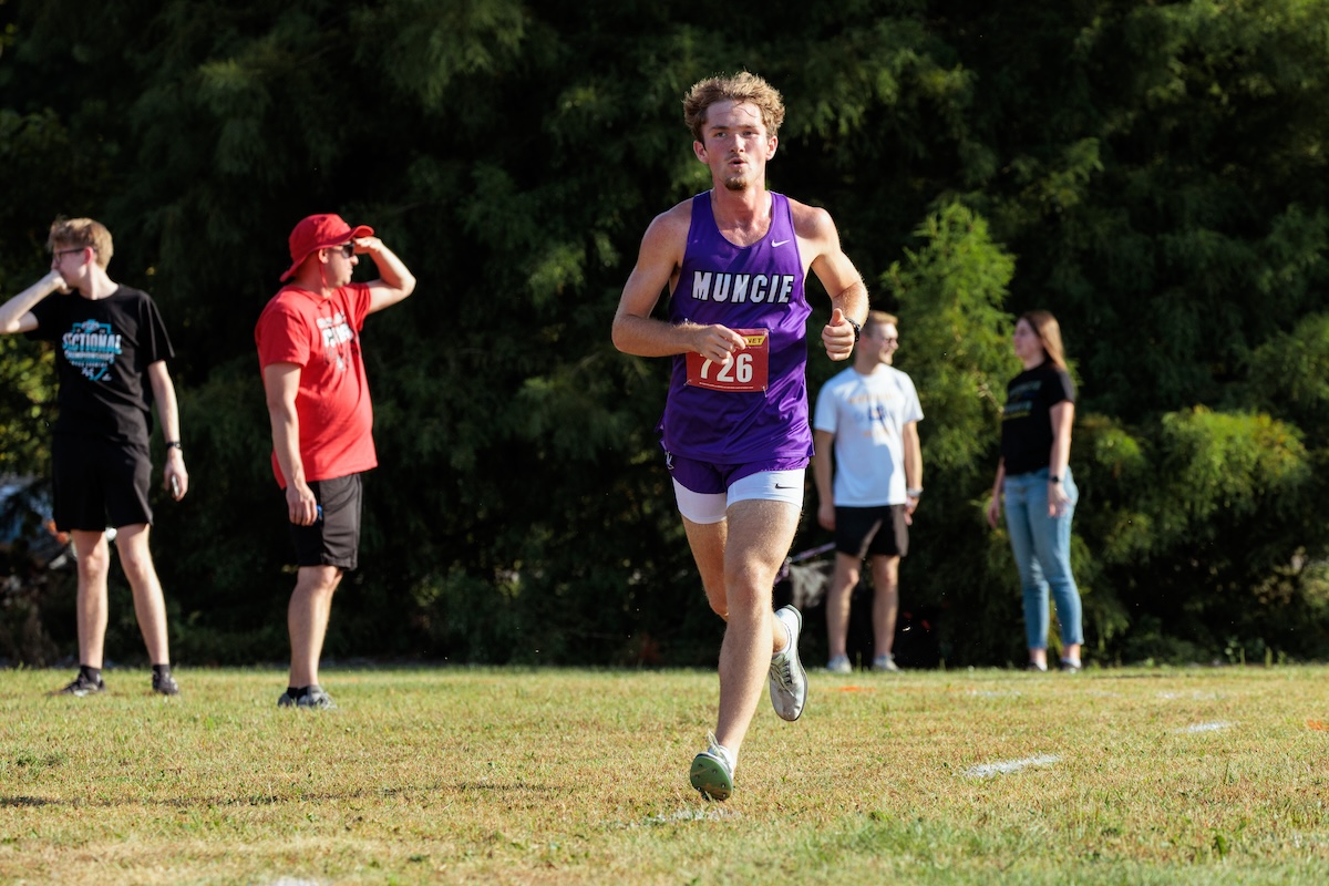 A young male cross-country runner with curly hair is captured mid-stride during a race. He is wearing a purple singlet with "MUNCIE" in white letters across the chest and a red race bib numbered "726." He is running on a grassy course toward the camera, with a focused expression. In the blurred background, several spectators stand near a line of dense green trees, including one man in a bright red shirt and hat shielding his eyes from the sun.