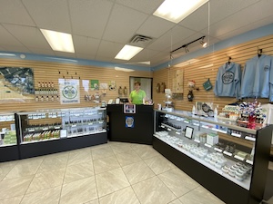 An interior shot of a well-lit retail store, likely a boutique or gift shop. A staff member in a bright green shirt stands behind a black L-shaped service counter in the center. The shop features glass display cases filled with small items, light wood-slat walls holding merchandise like apparel and accessories, and a neutral tiled floor. A circular green logo is visible on the wall behind the counter.
