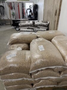 An indoor storage area or retail space featuring stacks of large clear bags filled with grain. In the background, additional branded feed bags labeled "Elite Nutrition" are stacked against a white wall. The room also contains various pieces of equipment, including a cart and what appears to be harness or rope gear hanging from a slatwall, suggesting a farm supply or animal nutrition store.