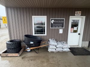 The storefront of Master Key Sales, a business with brown vertical siding and a white door. A black and white sign features a horse head logo and the tagline "Unlock the Natural Potential," noting they specialize in non-GMO and natural products. Outside on the concrete porch, several items are displayed for sale: a stack of large tractor tires, a black wheelbarrow with wooden handles holding two large plastic tubs, and a pallet of stacked grey bags. A small "Farm Fresh Eggs for Sale" sign is posted above the door.
