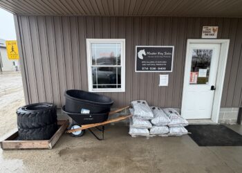 The storefront of Master Key Sales, a business with brown vertical siding and a white door. A black and white sign features a horse head logo and the tagline "Unlock the Natural Potential," noting they specialize in non-GMO and natural products. Outside on the concrete porch, several items are displayed for sale: a stack of large tractor tires, a black wheelbarrow with wooden handles holding two large plastic tubs, and a pallet of stacked grey bags. A small "Farm Fresh Eggs for Sale" sign is posted above the door.