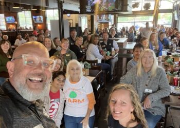 A high-angle selfie capturing a large, cheerful group of people gathered inside a modern restaurant and brewery, likely Bier Brewery Noblesville. In the foreground, a man with a grey beard, glasses, and a wide smile looks into the camera. Near him, several women are visible, including one wearing a white Teter Organic Farm t-shirt with a green cabbage logo. The background is filled with more smiling attendees seated at long wooden tables. The industrial-style space features high ceilings, exposed black beams, multiple TV screens, and large glass garage doors that let in natural light.