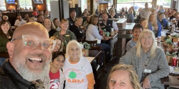 A high-angle selfie capturing a large, cheerful group of people gathered inside a modern restaurant and brewery, likely Bier Brewery Noblesville. In the foreground, a man with a grey beard, glasses, and a wide smile looks into the camera. Near him, several women are visible, including one wearing a white Teter Organic Farm t-shirt with a green cabbage logo. The background is filled with more smiling attendees seated at long wooden tables. The industrial-style space features high ceilings, exposed black beams, multiple TV screens, and large glass garage doors that let in natural light.