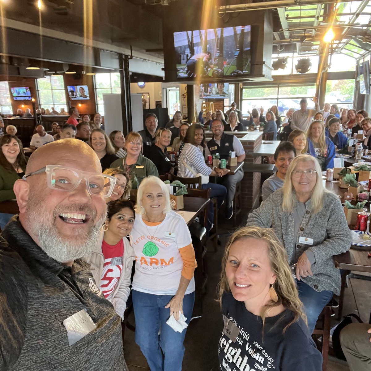 A high-angle selfie capturing a large, cheerful group of people gathered inside a modern restaurant and brewery, likely Bier Brewery Noblesville. In the foreground, a man with a grey beard, glasses, and a wide smile looks into the camera. Near him, several women are visible, including one wearing a white Teter Organic Farm t-shirt with a green cabbage logo. The background is filled with more smiling attendees seated at long wooden tables. The industrial-style space features high ceilings, exposed black beams, multiple TV screens, and large glass garage doors that let in natural light.