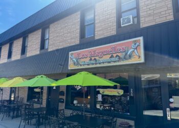 A street-level exterior view of Los Maguelles Mexican Restaurant in Fortville, Indiana. The restaurant is housed in a two-story tan brick building with dark metal siding on the right side. A large rectangular sign above the storefront features the restaurant's name in red cursive and includes illustrations of men harvesting agave plants. On the sidewalk, three bright lime-green patio umbrellas shade black metal tables and chairs, ready for outdoor dining. The glass front windows and a black door with the address "222" are visible beneath a dark grey metal awning.