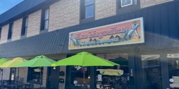 A street-level exterior view of Los Maguelles Mexican Restaurant in Fortville, Indiana. The restaurant is housed in a two-story tan brick building with dark metal siding on the right side. A large rectangular sign above the storefront features the restaurant's name in red cursive and includes illustrations of men harvesting agave plants. On the sidewalk, three bright lime-green patio umbrellas shade black metal tables and chairs, ready for outdoor dining. The glass front windows and a black door with the address "222" are visible beneath a dark grey metal awning.